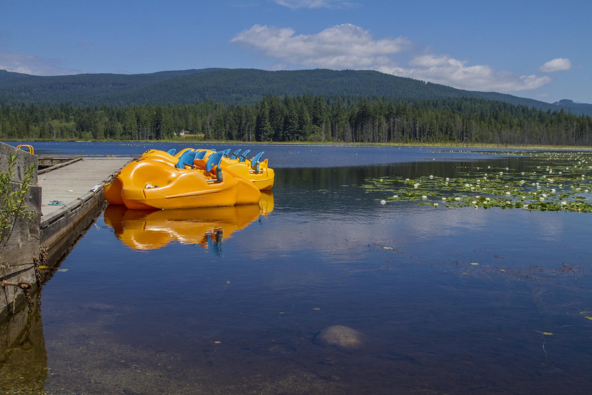 A view of a lake from dock level. Several yellow paddle boats line the doc and mountains rise in the background.