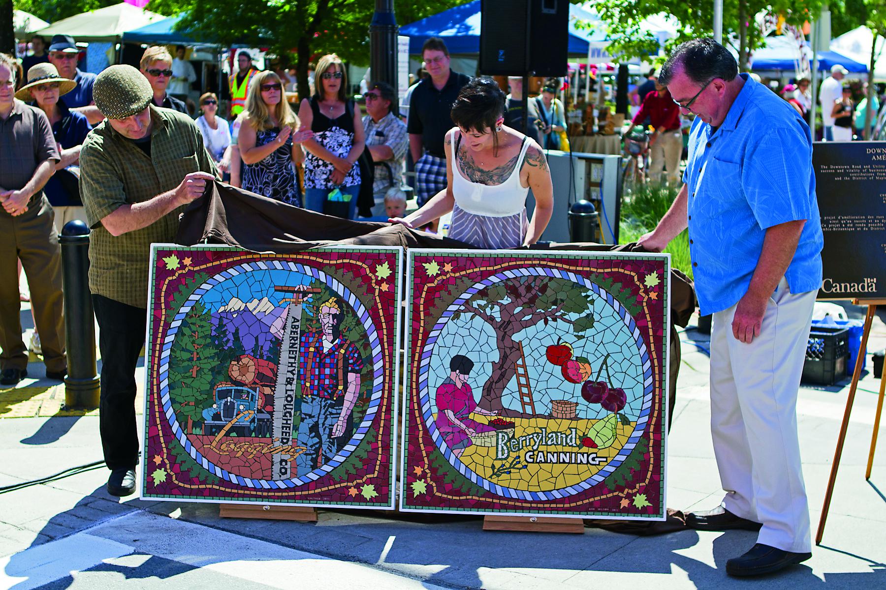 A crowd watches as three people display two tile mosaics. One depicts a man with logging equipment, while the other shows an androgynous worker sitting under a tree selecting and next to various fruit. White lettering proclaims "Berryland Canning."