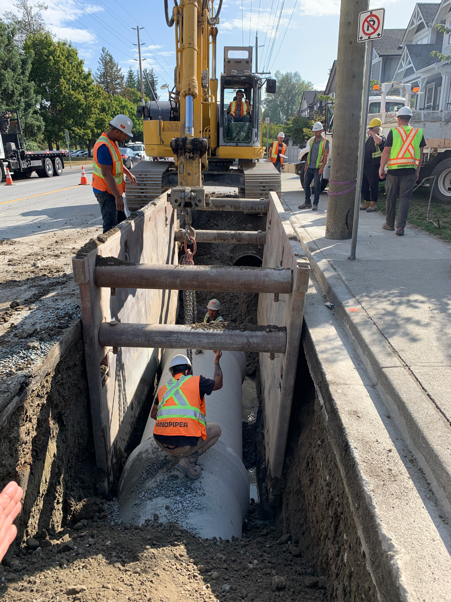 A crew of workers wearing high-visibility vests set a section of pipe into a construction canal next to the sidewalk.