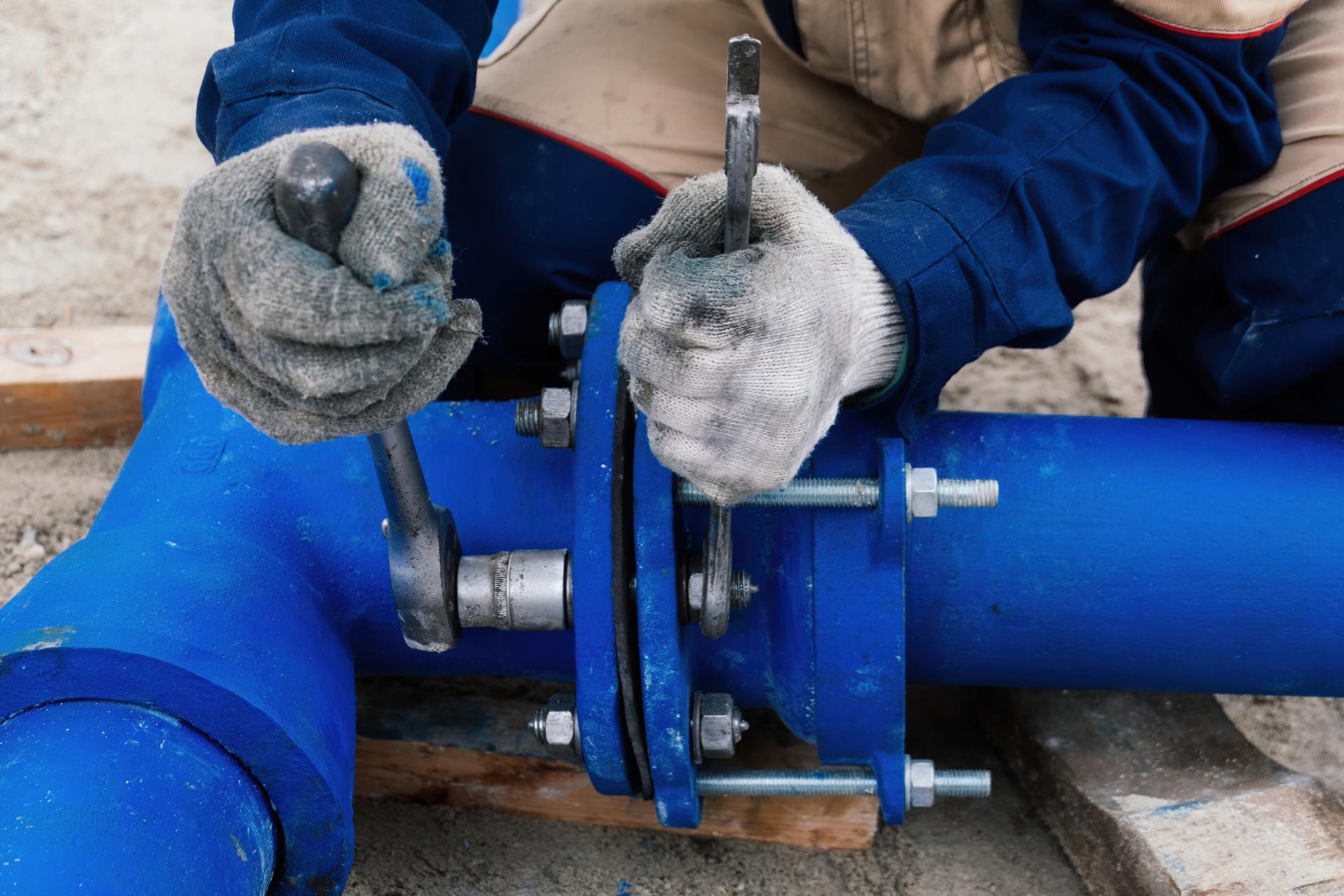 large blue water main pipe being adjusted by a tradesperson kneeling over it, wearing gloves and adjusting it with two wrenches