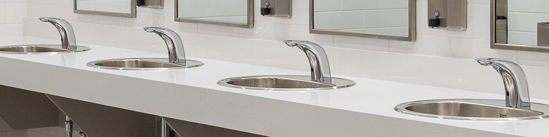 A row of four silver sinks sit waiting to be used in a public washroom.