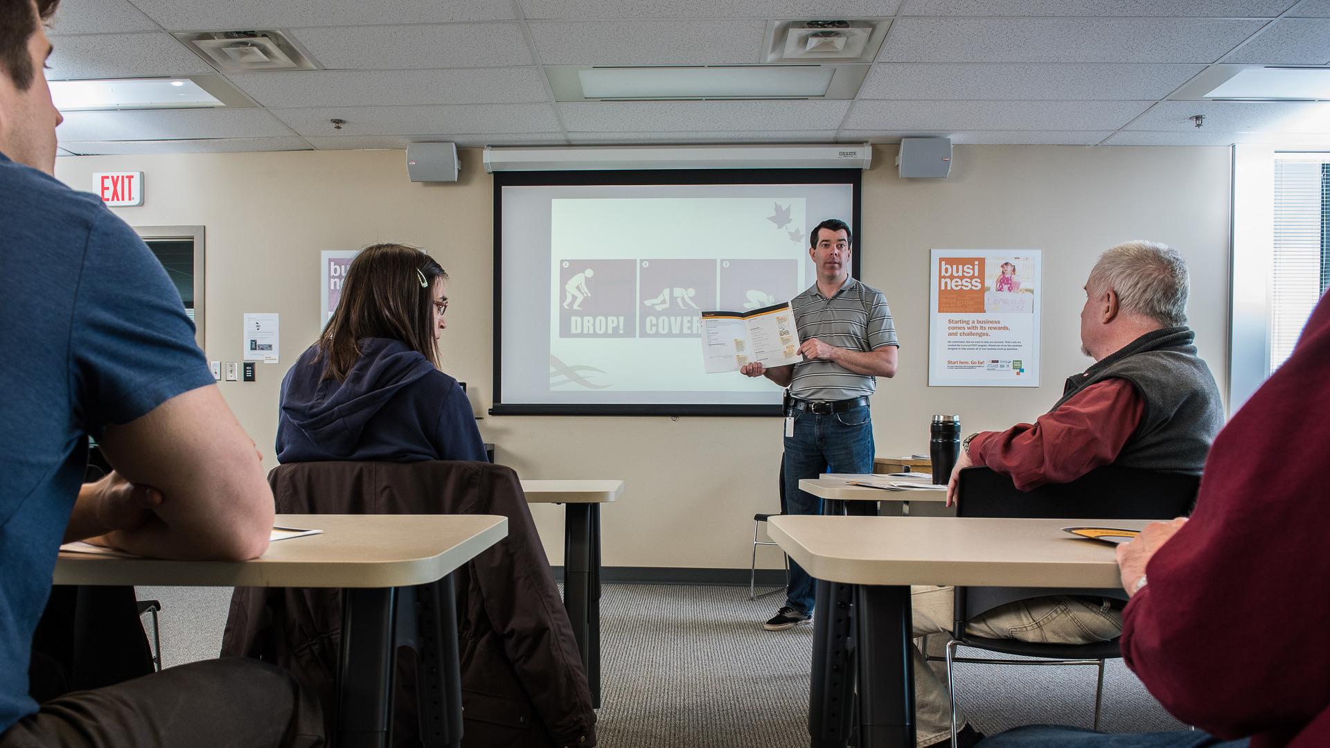 A man stands at the front of a classroom with a book open, and earthquake instructions on a screen behind him, teaching volunteers.