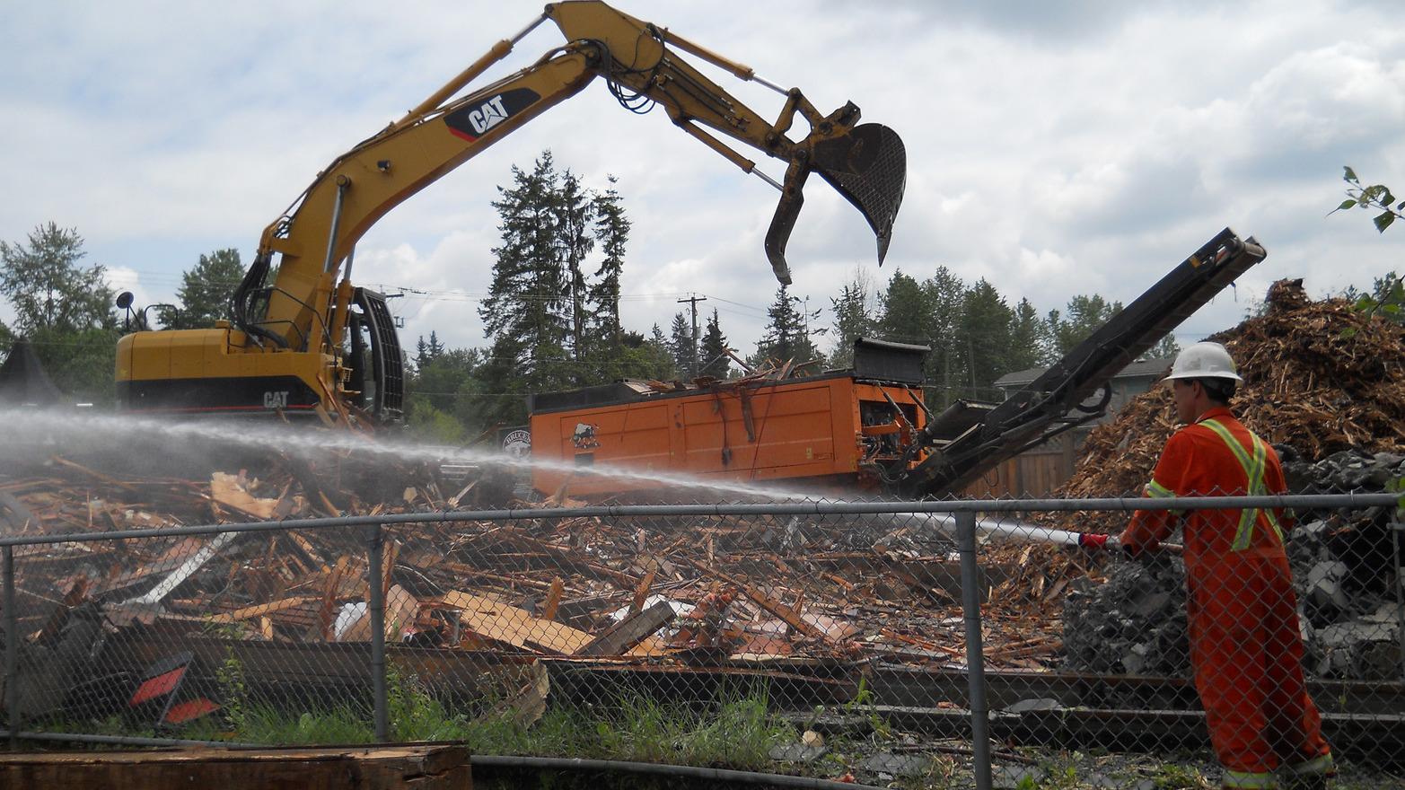An excavator finishes demolishing a building while a worker houses it down with water to prevent dust from entering the air.