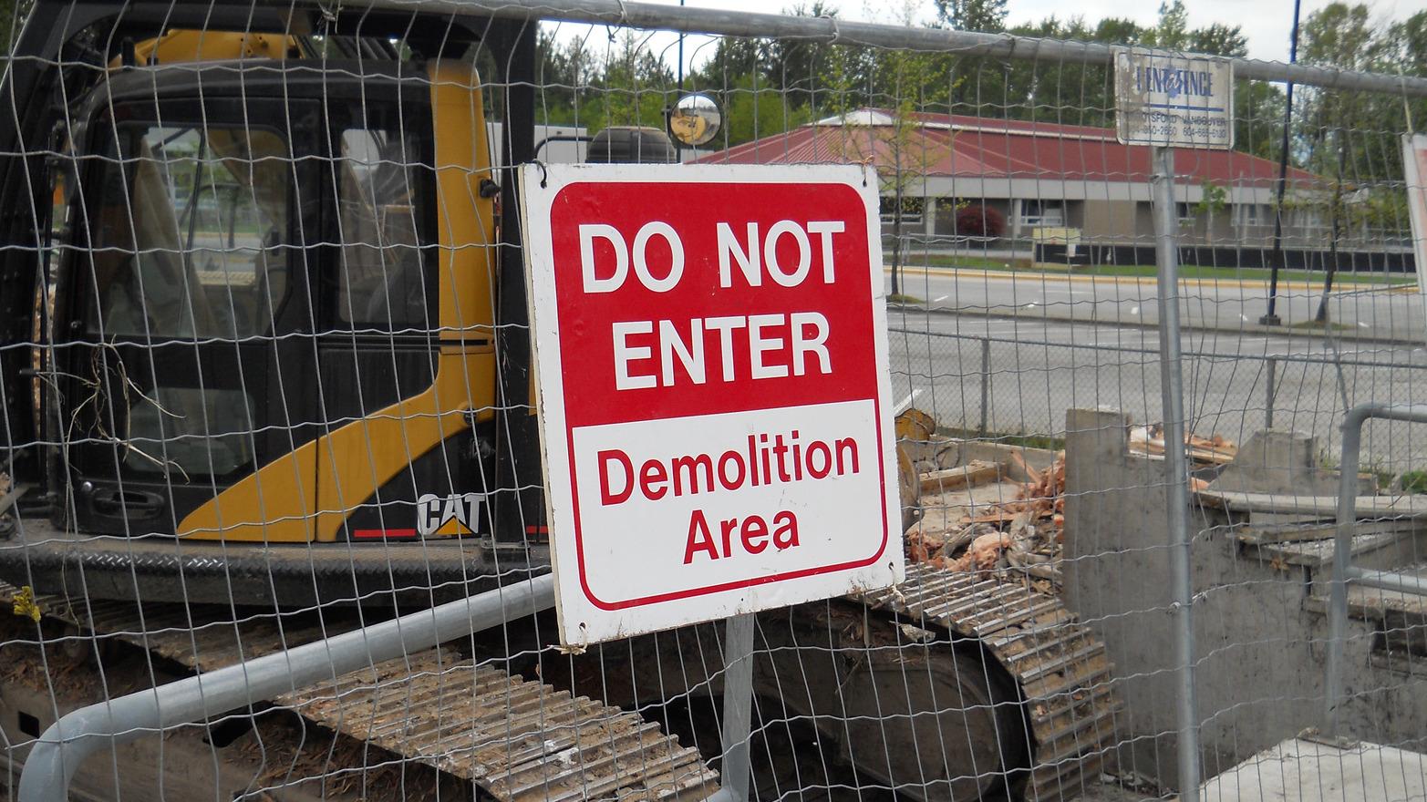 A sign saying "Do not enter, Demolition Area" is affixed to the fence next to an excavator at a demolition site.