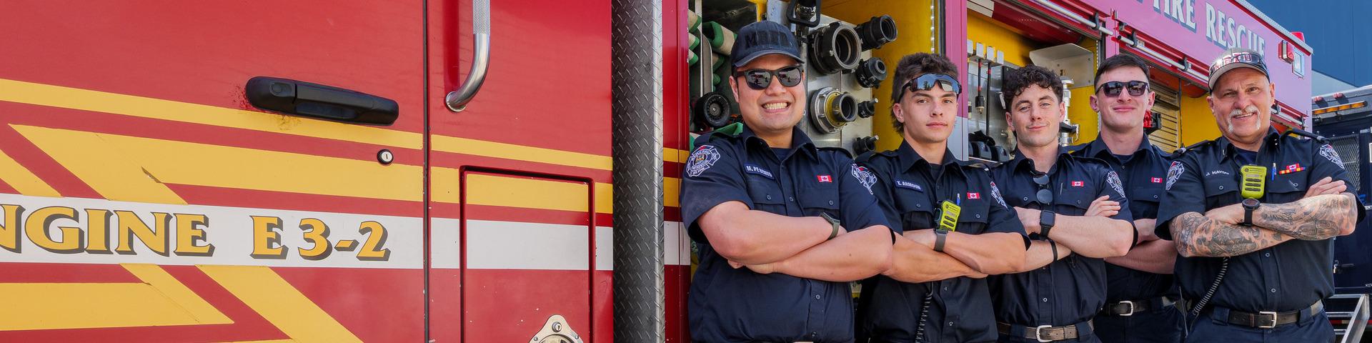 Five firefighters stand smiling with their arms crossed next to a fire truck.