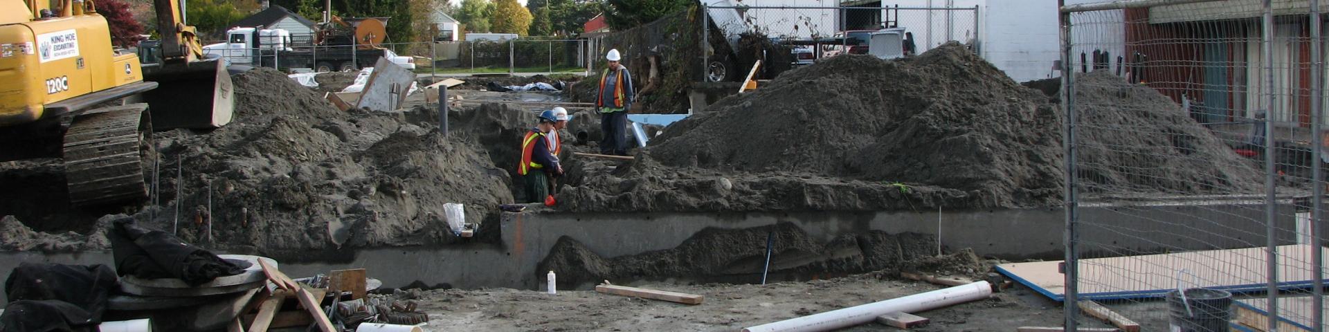 Workers in safety gear traverse a construction site where the foundation has been dug up.