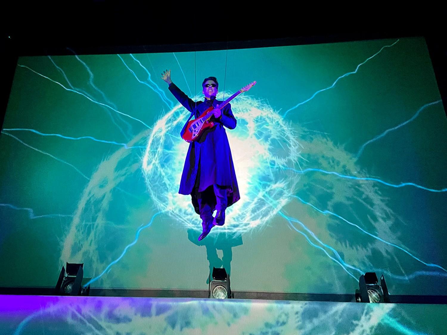 A man lit in purple rises above a stage to play a guitar while cyan lightning dances on the stage backing behind him.