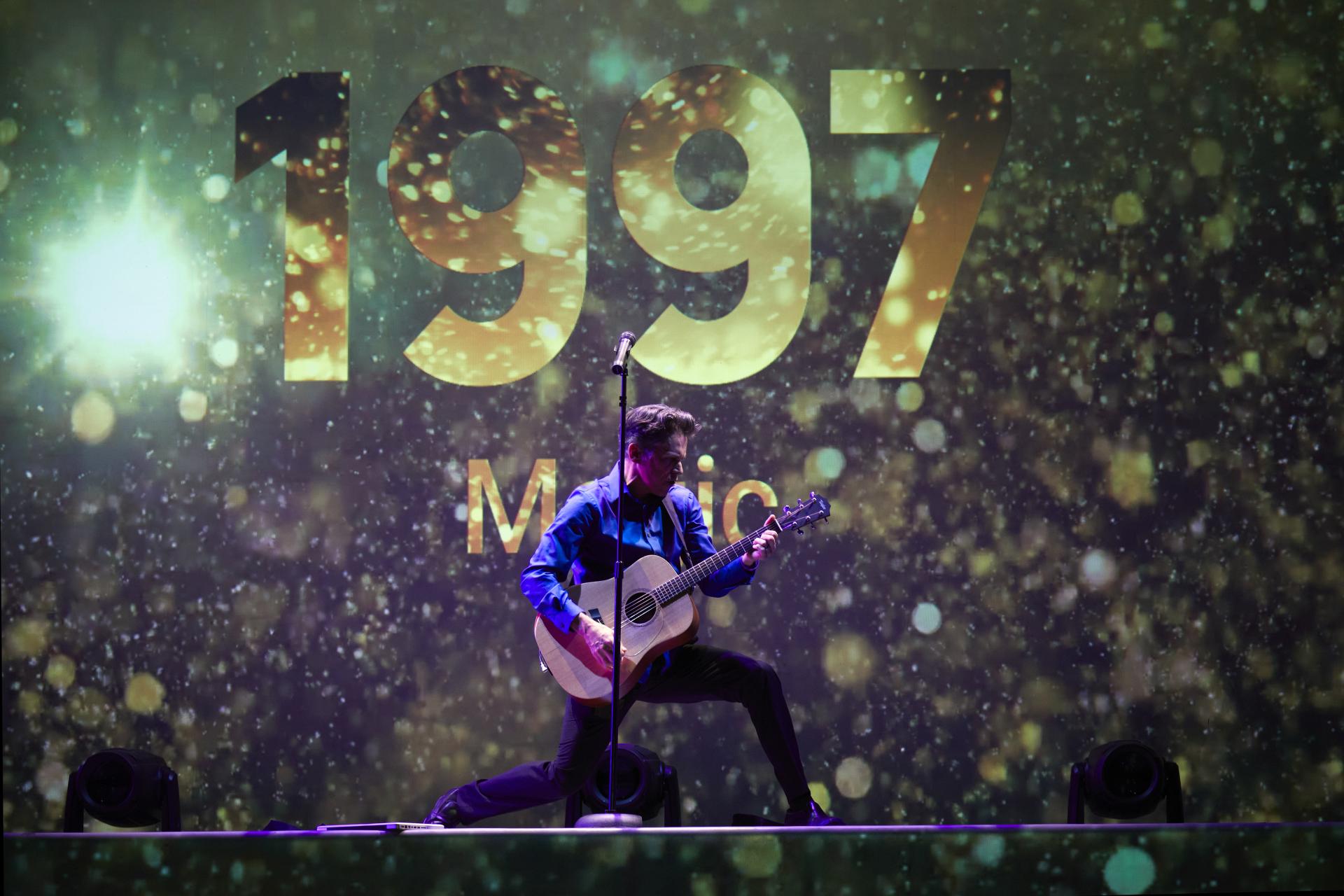 A man playing a guitar kneels dramatically before a screen that proclaims 1997 in gold numerals.