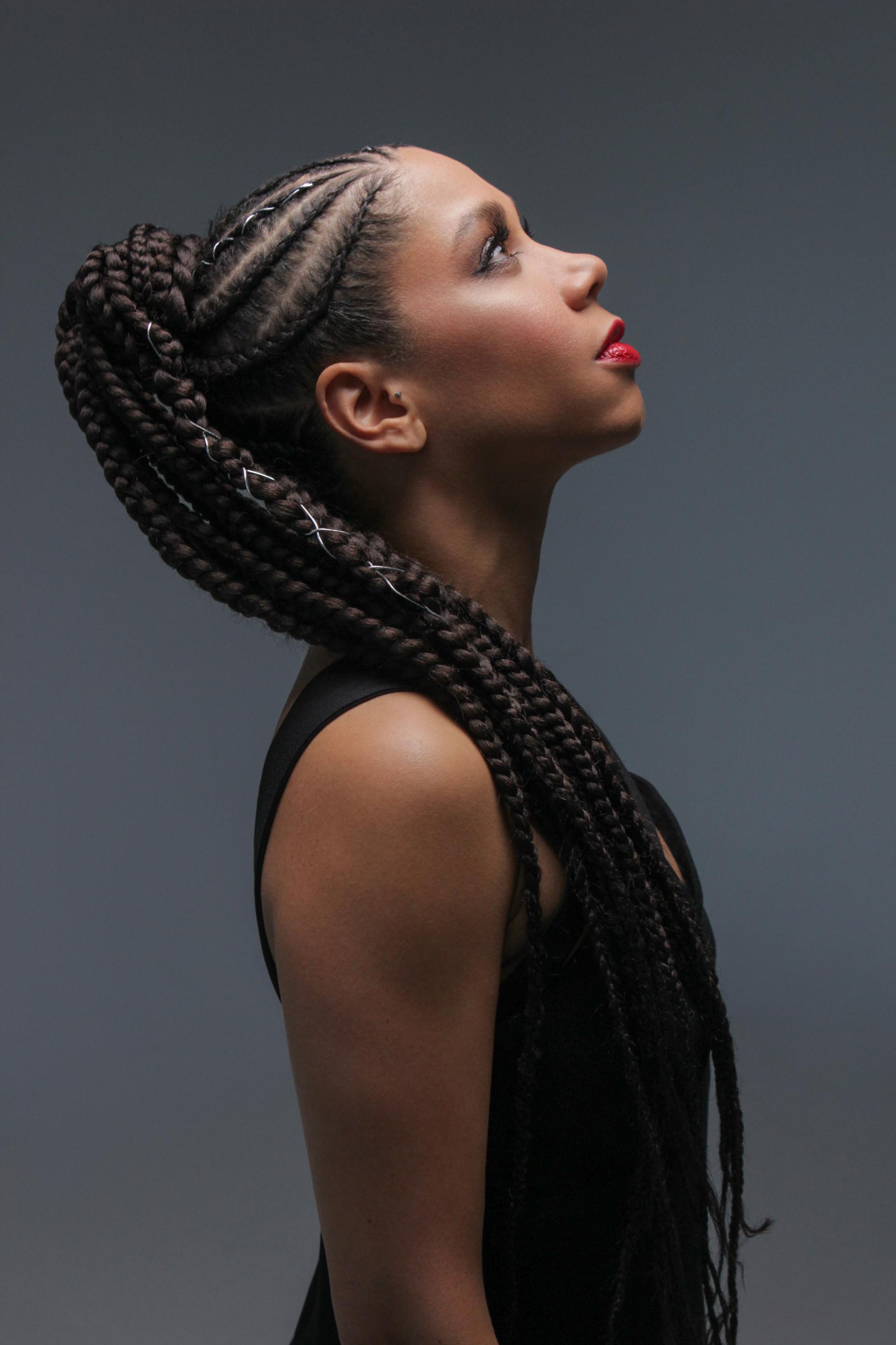 Tina Leon, wearing thick braids looks up towards the light against a striking grey background.