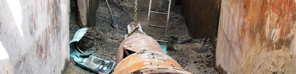 A large rusty pipe at the bottom of a cut channel, with a ladder leading upwards.