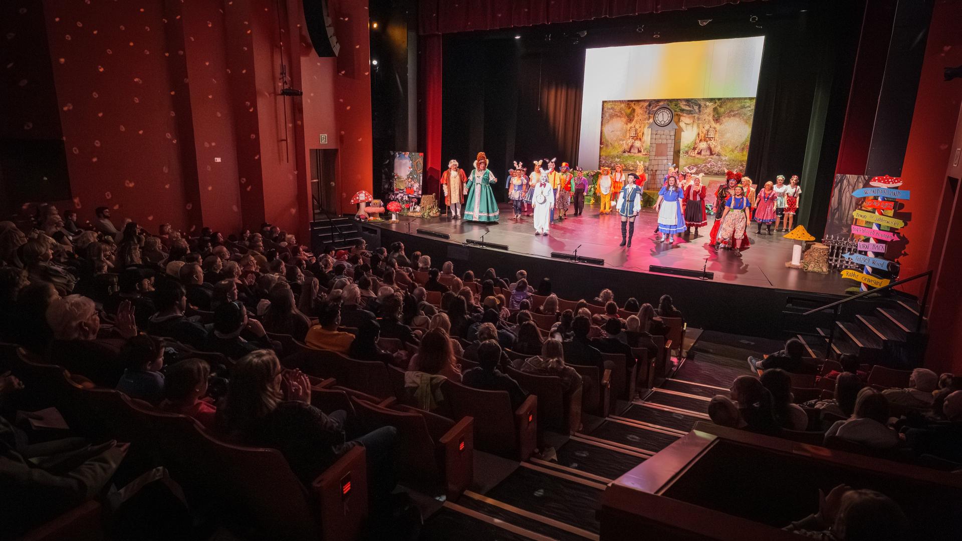 The cast of a colourful rendition of "Alice in Wonderland" stands before a seated crowd at the ACT Arts Centre Main Stage Theatre.