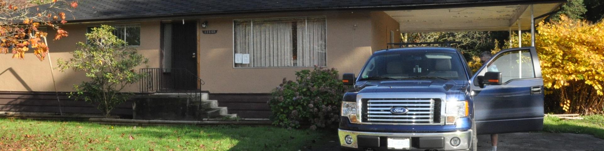 A large truck rests just out from under the cover of a carport attached to a brown house.