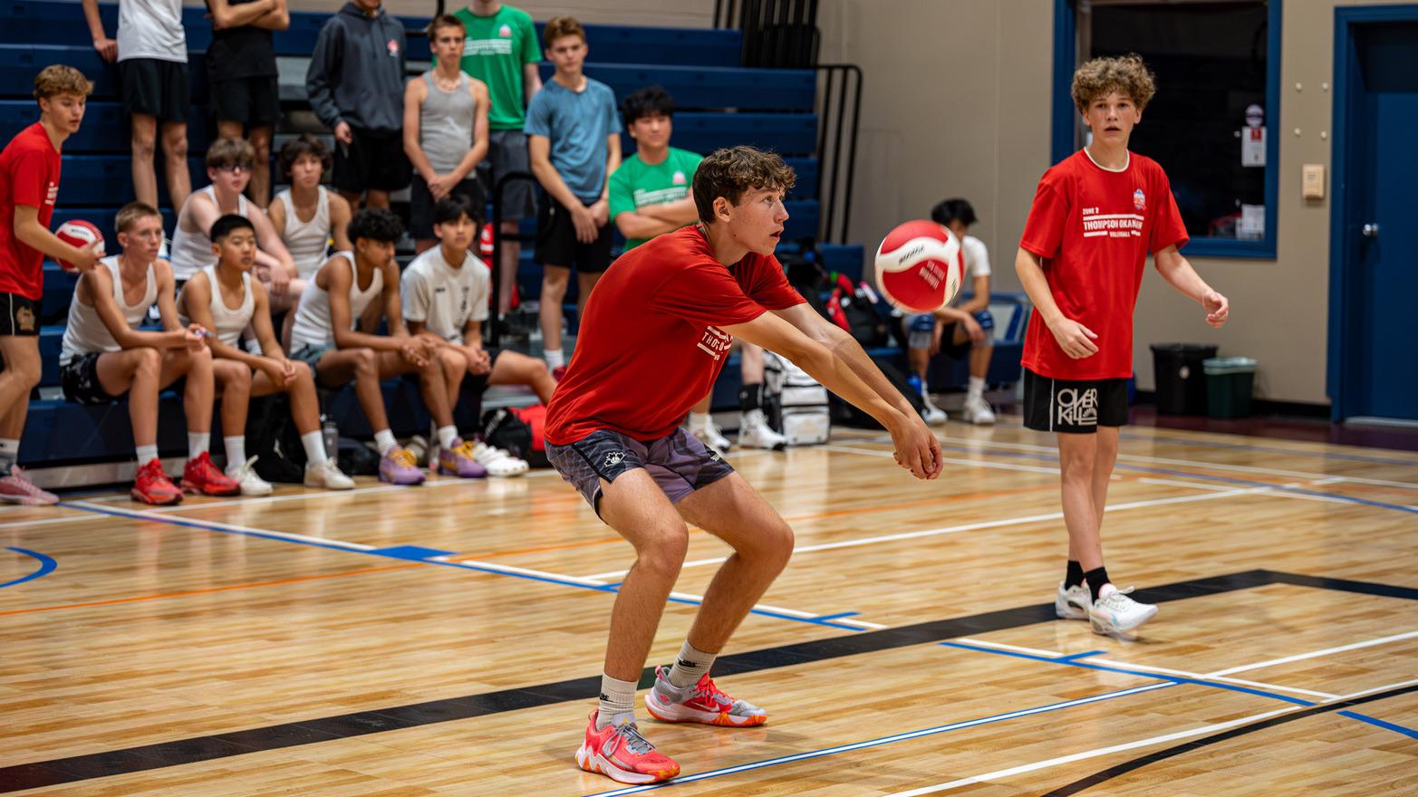 A boy in an indoor sports court prepares to return a flying volleyball while his teammate and a crowd seated on bleaches watches.