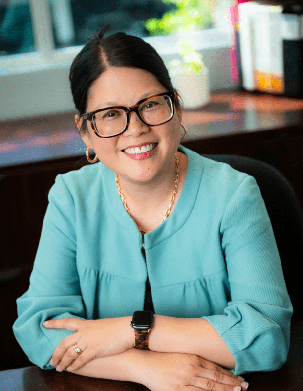 Shauna Harper smiles at the camera from inside an office, wearing a light blue blouse and dark, thick-rimmed glasses.