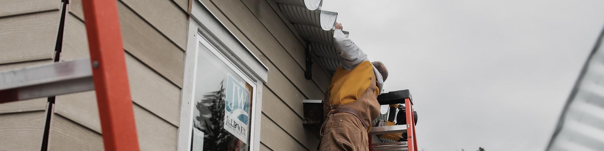 A man stands next to a ladder, reaching up to work on the shiny silver tiles of a roof.