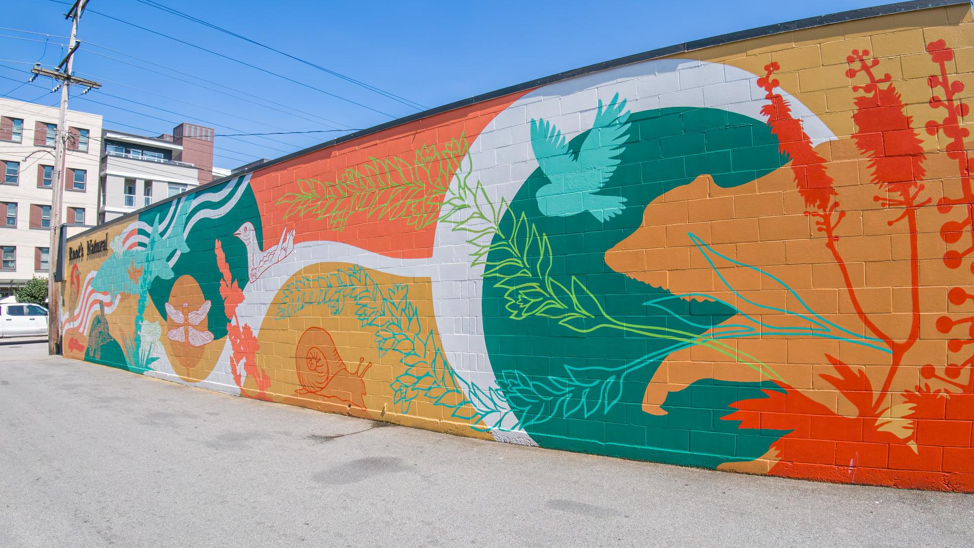 A vibrant mural in reds, teal and oranges depicts native species, such as a heron, dragonfly, snail, and owl alongside plant imagry.